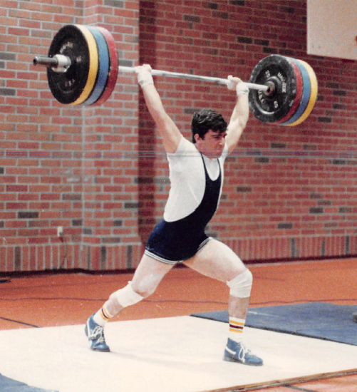 The author, weightlifting in a gymnasium circa 1984. The large weights attached to a barbell and being held overhead are 145kg or roughly 320lbs.